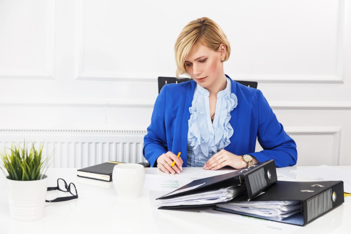 Žena poslovno obučena sjedi za bijelim radnim stolom na kojem su registratori, rokovnik, naočare i biljka